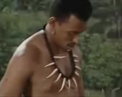 A fucker with a necklace of animal teeth and a beard is looking away from the camera.