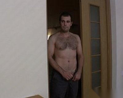 A bearded fucker without a shirt standing in a hallway.