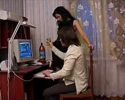 A person is sitting at a computer. Another person is standing next to two chairs.
